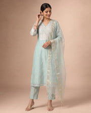 A woman modeling a light blue-green traditional Indian salwar kameez suit with white embroidery, matching trousers, and a sheer white dupatta, set against a solid beige studio background.