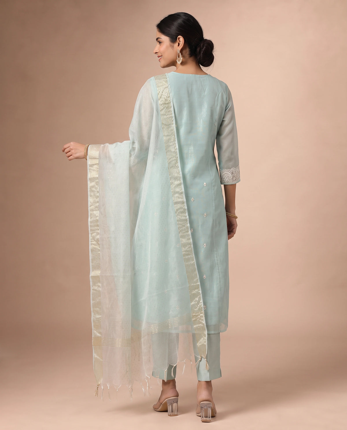 A woman modeling a light blue traditional outfit with a white dupatta featuring gold borders, shown from the back against a solid beige studio background.