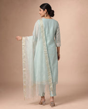 A woman modeling a light blue traditional outfit with a white dupatta featuring gold borders, shown from the back against a solid beige studio background.
