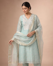 A woman models a traditional light blue salwar kameez suit with a matching sheer dupatta. The kurta features white embroidery on the chest and small gold floral motifs, paired with light blue trousers.