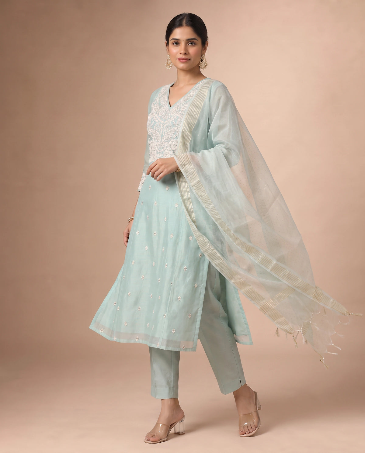 A woman models a light mint green ethnic salwar kameez suit with white embroidery, matching pants, and a sheer white dupatta, set against a solid beige studio background.