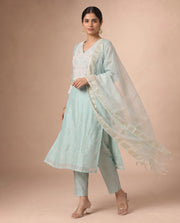 A woman models a light mint green ethnic salwar kameez suit with white embroidery, matching pants, and a sheer white dupatta, set against a solid beige studio background.