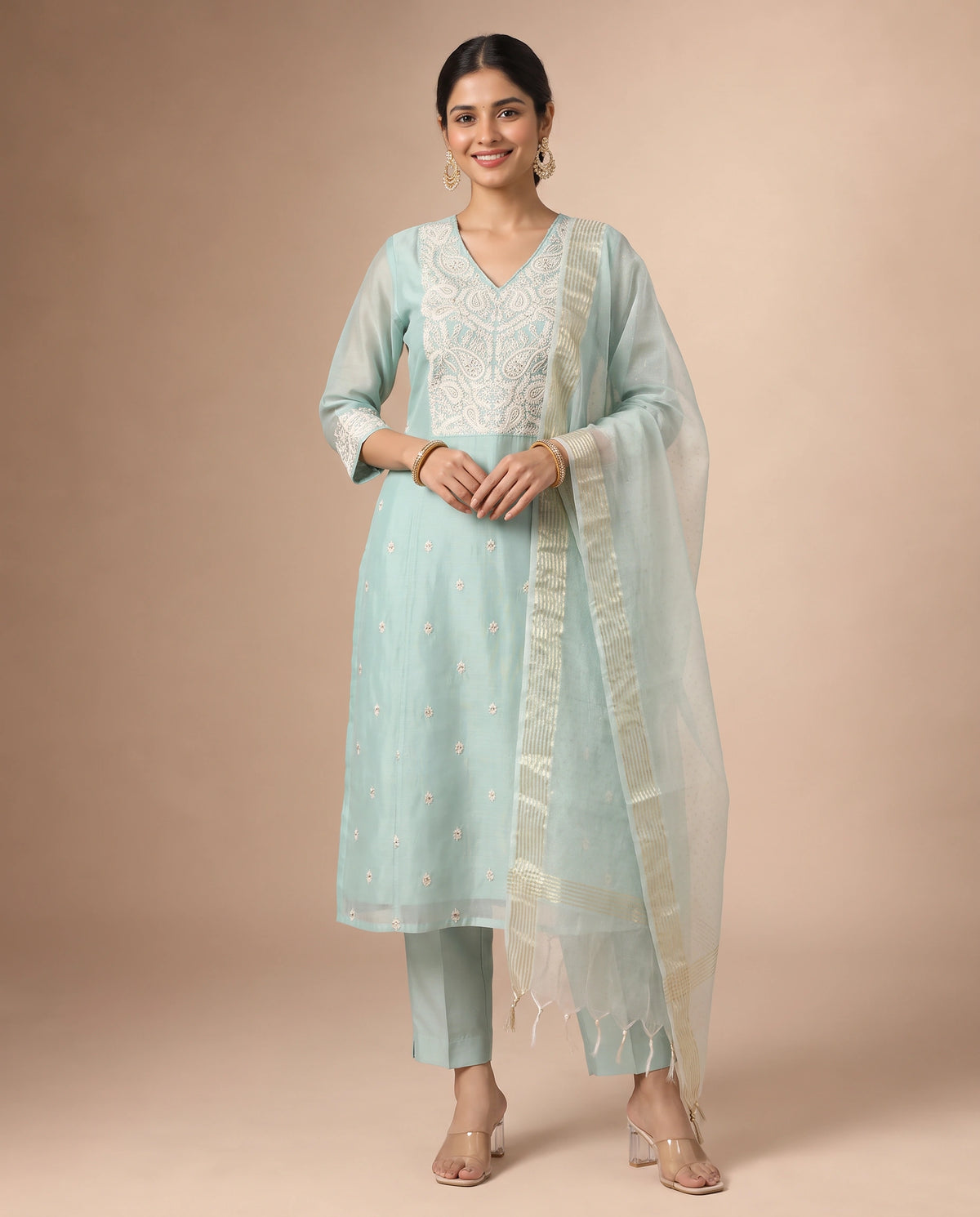 A woman modeling a light blue-green traditional Indian salwar kameez suit with white embroidery, matching trousers, and a sheer dupatta, set against a solid beige studio background.