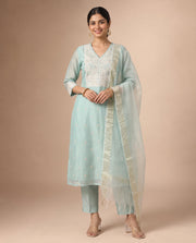A woman modeling a light blue-green traditional Indian salwar kameez suit with white embroidery, matching trousers, and a sheer dupatta, set against a solid beige studio background.