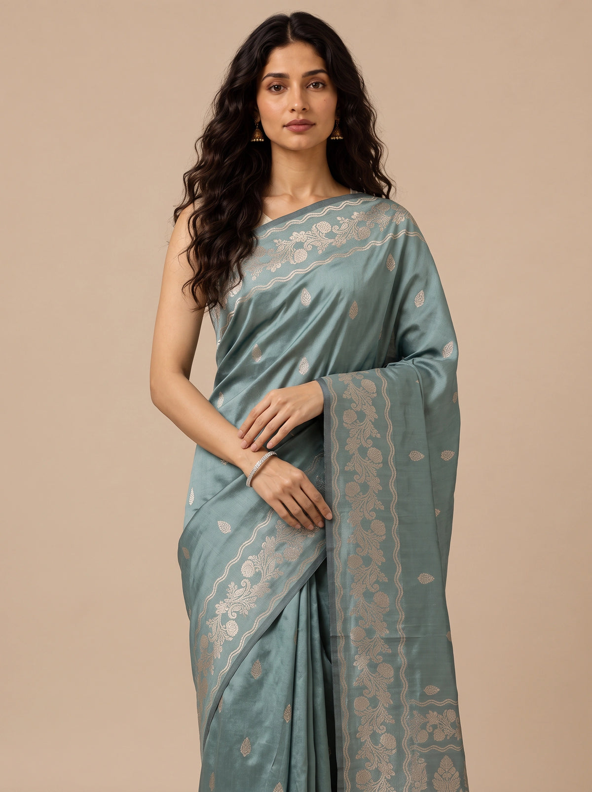 A woman modeling a teal silk saree with intricate gold zari floral patterns and borders, set against a solid beige studio background.