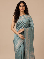A woman modeling a teal silk saree with intricate gold zari floral patterns and borders, set against a solid beige studio background.