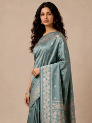 A woman modeling a teal saree with intricate silver floral border and small metallic motifs, set against a solid beige studio background.