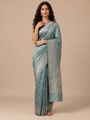 A woman modeling a Teal silk saree with metallic silver zari floral patterns and a wide border, set against a solid beige studio background.