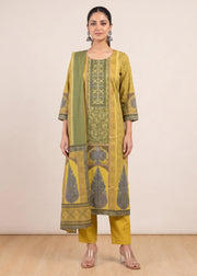 A woman modeling a traditional Indian salwar kameez suit set consisting of a mustard yellow kurta with green and gray printed patterns, matching yellow trousers, and a green dupatta, set against a plain beige studio background.