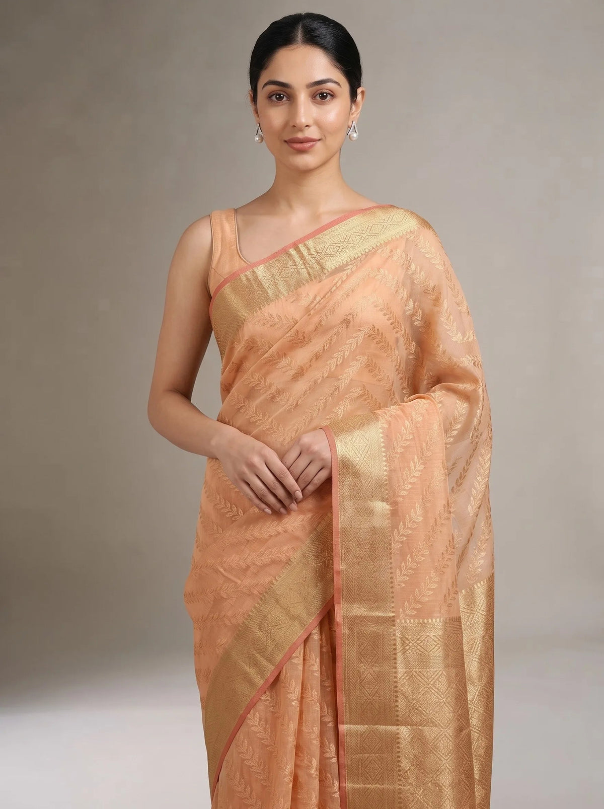 A woman modeling a peach-colored silk saree with a wide gold zari border and a subtle woven pattern, set against a neutral gray studio background.